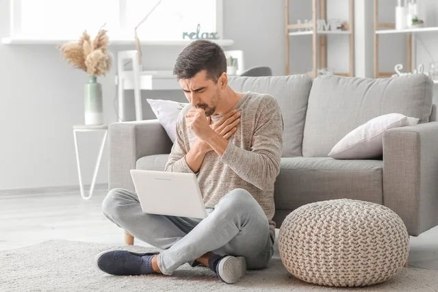 Hombre tosiendo sentado en un sillón con una computadora