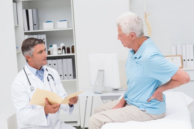 older male patient with hand on his lower back talking to doctor seated at the bedside